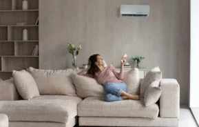 Mulher relaxando em sofá confortável, usando controle para ar-condicionado e em ambiente modernamente decorado e climatizado.