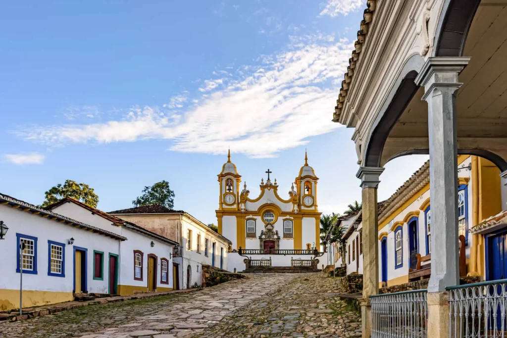 Imagem da paisagem urbana de Tiradentes mostra rua de paralelepípedos, construções coloniais e igreja ao fundo para ilustrar matéria sobre quais as cidades históricas de Minas Gerais