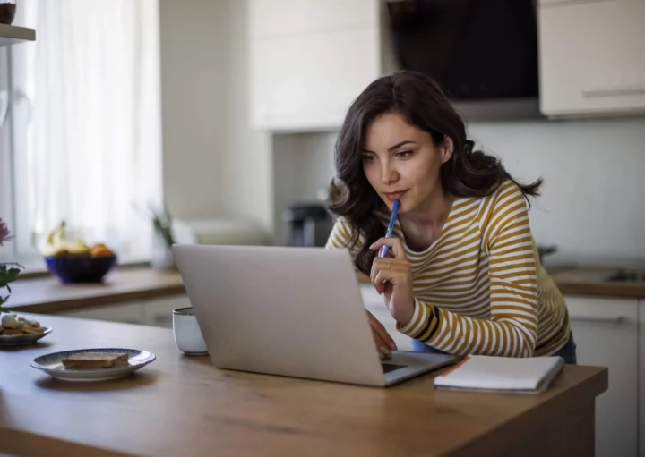 Imagem de uma mulher usando o notebook na bancada da cozinha de casa enquanto aponta uma caneta azul para o queixo para ilustrar matéria sobre qual o reajuste de aluguel permitido por lei