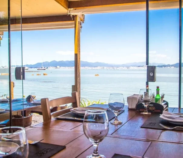Foto que ilustra matéria sobre os melhores restaurantes em Florianópolis mostra o ambiente interno do Rosso Restaurante com uma vista para o mar através de uma grande janela de vidro (Foto: Divulgação)