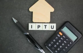 Imagem das iniciais de “IPTU” em dados em cima de uma bancada ao lado de uma caneta, uma calculadora e uma casa de madeira em tamanho miniatura para ilustrar matéria sobre cidades com IPTU mais caro do Brasil