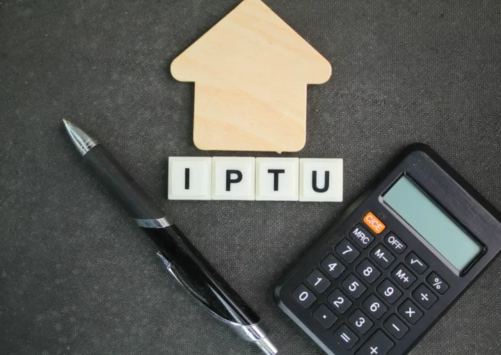 Imagem das iniciais de “IPTU” em dados em cima de uma bancada ao lado de uma caneta, uma calculadora e uma casa de madeira em tamanho miniatura para ilustrar matéria sobre cidades com IPTU mais caro do Brasil