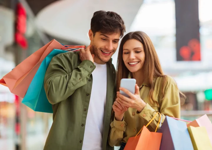 Imagem de um casal em um shopping carregando algumas sacolas enquanto a mulher mostra algo no celular ao homem para ilustrar matéria sobre o Shopping Eldorado