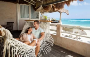 Imagem de um casal feliz formado por um homem e uma mulher deitados em uma rede que está na varanda de uma casa em frente a praia para ilustrar matéria sobre imóveis de veraneio