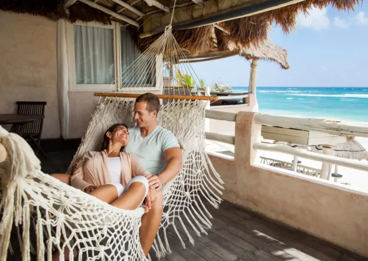Imagem de um casal feliz formado por um homem e uma mulher deitados em uma rede que está na varanda de uma casa em frente a praia para ilustrar matéria sobre imóveis de veraneio