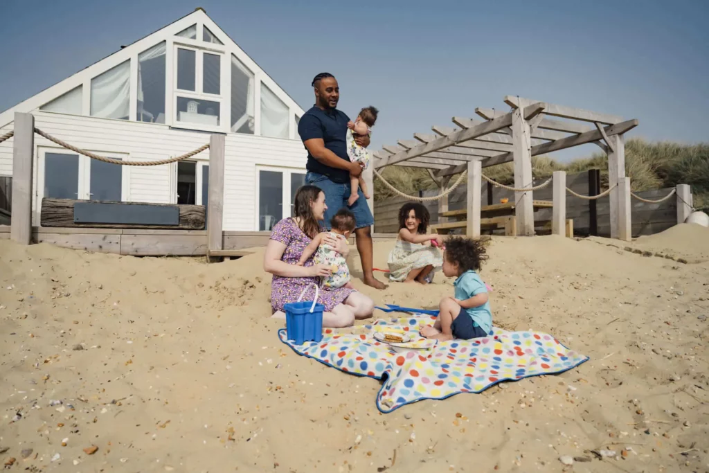 Imagem de uma família formada por um homem, uma mulher e quatro crianças brincando na areia da praia em frente a um imóvel para ilustrar matéria sobre o que significa uma casa de veraneio