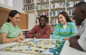 Imagem de pessoas amigos ao redor de uma mesa jogando um jogo de tabuleiro com uma prateleira com vários livros e jogos atrás para ilustrar matéria sobre bar com jogo de tabuleiro