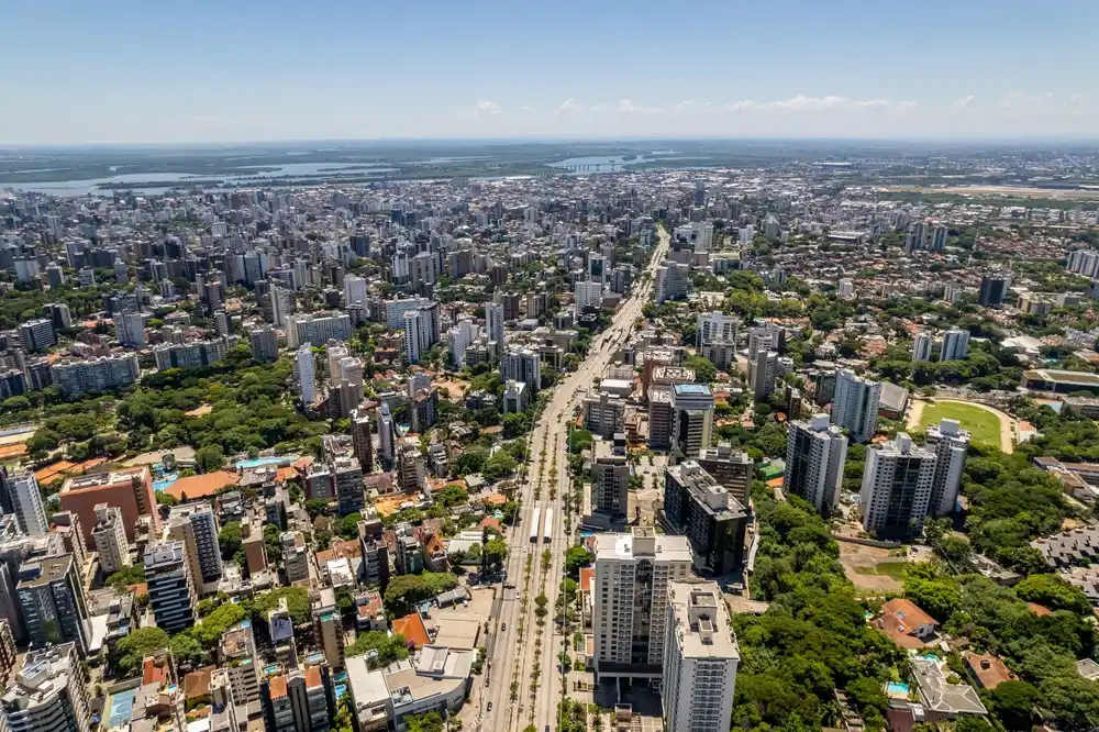 Imagem da vista aérea de Porto Alegre mostra cenário urbano da cidade com prédios, casas, vegetação e uma avenida em um dia de céu azul