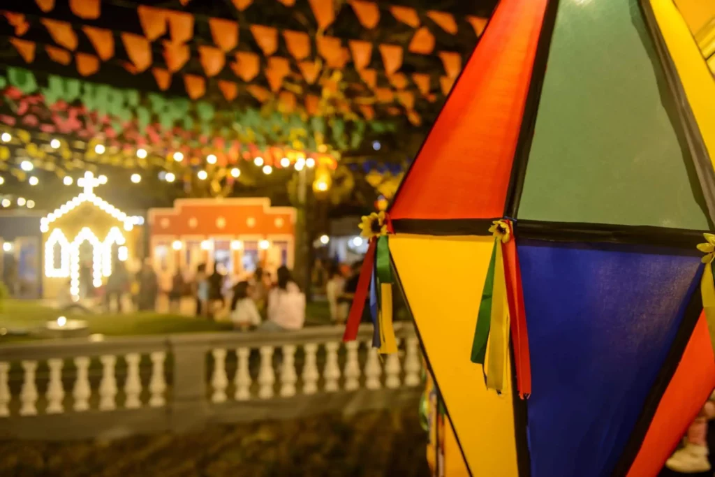 Imagem de bandeiras coloridas e balão decorativo, além de estrutura de festa e pessoas ao fundo, para ilustrar matéria sobre festa junina em São Paulo