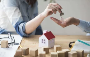 Imagem de uma pessoa entregando as chaves de casa para outra pessoa próximo a uma mesa onde estão uma casa em tamanho miniatura, alguns blocos de madeira, um óculos, um caderno e uma caneta para ilustrar matéria sobre o imposto sobre compra e venda de imóvel