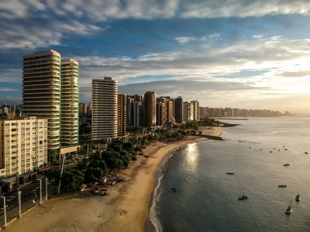 Vista aérea da Praia do Mucuripe, em Fortaleza, mostra mar, faixa de areia e prédios da orla durante o pôr do sol
