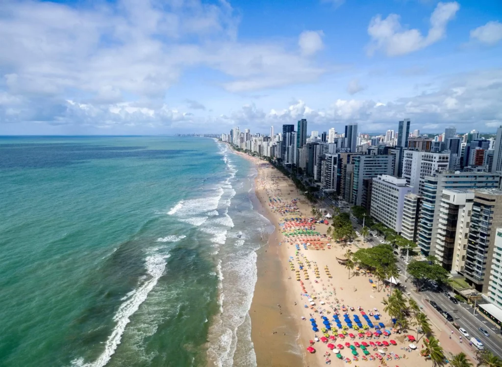 Imagem da vista aérea da Praia de Boa Viagem, em Recife, mostra orla, prédios, mar e turistas na areia em um dia de céu azul