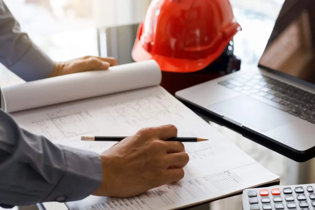 Imagem de um homem segurando um lápis para escrever em um projeto arquitetônico que está em cima da mesa em frente a um notebook para ilustrar matéria como como calcular o valor da averbação da construção