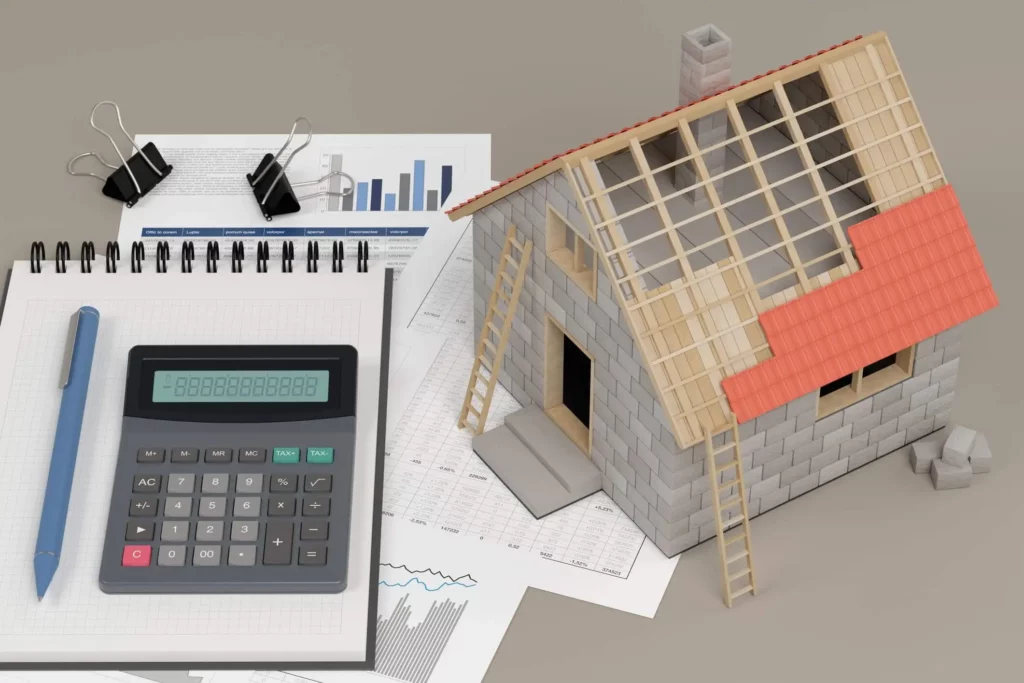 Imagem de uma casa de madeira em tamanho miniatura com o telhado inacabado em cima de uma mesa ao lado de papéis, uma caneta e uma calculadora para ilustrar matéria sobre como fazer orçamento de obra