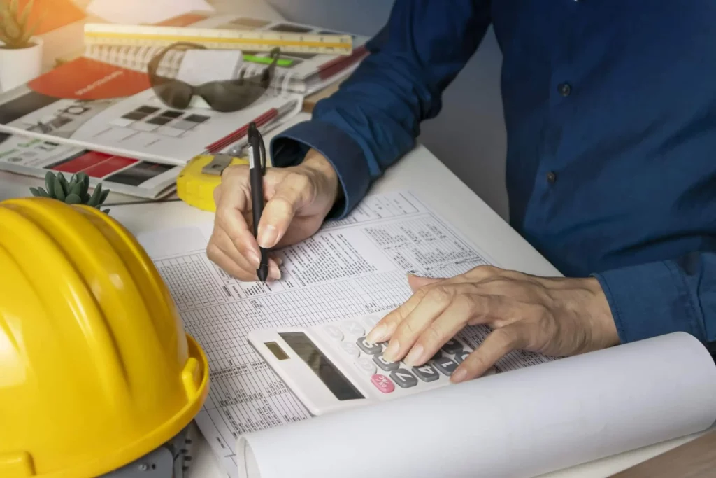Imagem de um homem escrevendo em uma planilha enquanto faz cálculos em uma calculadora que está em cima da mesa ao lado de um capacete de construção civil para ilustrar matéria sobre como preparar orçamentos de obras
