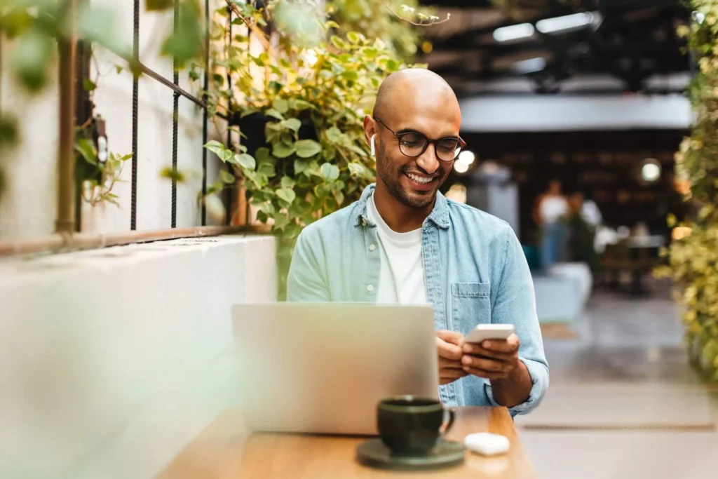 Imagem de um homem sorridente, usando óculos, sentado em uma mesa com um laptop e uma xícara de café, usando seu smartphone em um café bem iluminado e cheio de plantas