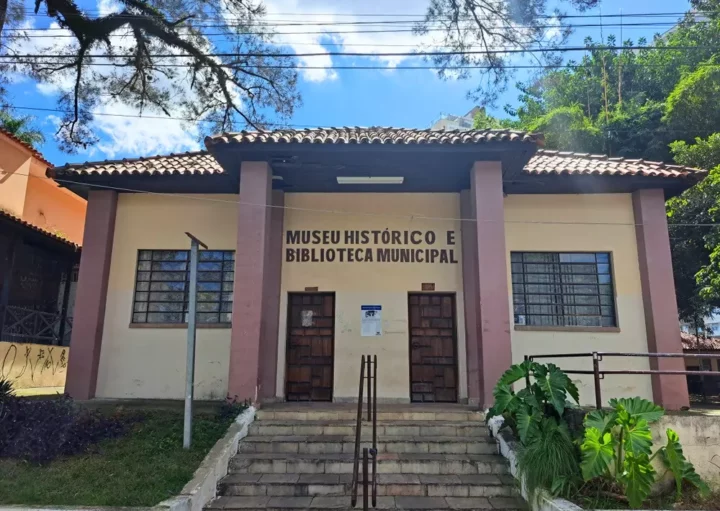 Um prédio bege com acabamento marrom e telhado de telhas, o Museu Histórico Municipal, é um dos notáveis Museus em Guarulhos, com duas portas, janelas gradeadas, degraus, vegetação e árvores sob um céu azul.