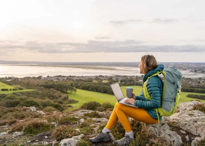 Imagem de uma pessoa usando uma mochila e roupas para atividades ao ar livre senta-se no topo de uma colina rochosa, usando um notebook e segurando uma xícara, com vista para uma paisagem cênica de campos, árvores, água e prédios distantes sob um céu nublado
