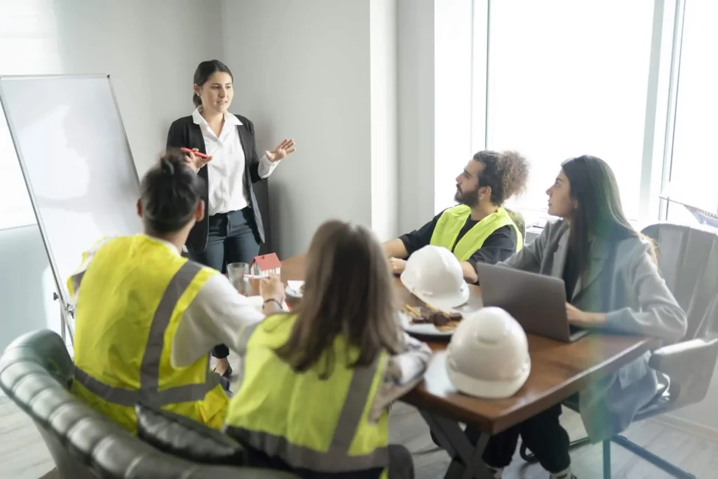 Imagem de profissionais da construção civil sentados ao redor de uma mesa escutando uma mulher que está ao lado de uma prancheta apresentado um projeto para ilustrar matéria sobre o que é orçamento de obra