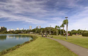 Um caminho pavimentado se curva no Parque Barigui, com palmeiras ao lado de um lago - uma das melhores opções de onde correr em Curitiba. Algumas sentam e relaxam na grama enquanto outras se exercitam na pista de corrida. Ao fundo prédios da cidade se erguem sob um céu azul parcialmente nublado.