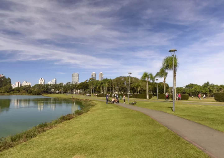 Um caminho pavimentado se curva no Parque Barigui, com palmeiras ao lado de um lago - uma das melhores opções de onde correr em Curitiba. Algumas sentam e relaxam na grama enquanto outras se exercitam na pista de corrida. Ao fundo prédios da cidade se erguem sob um céu azul parcialmente nublado.