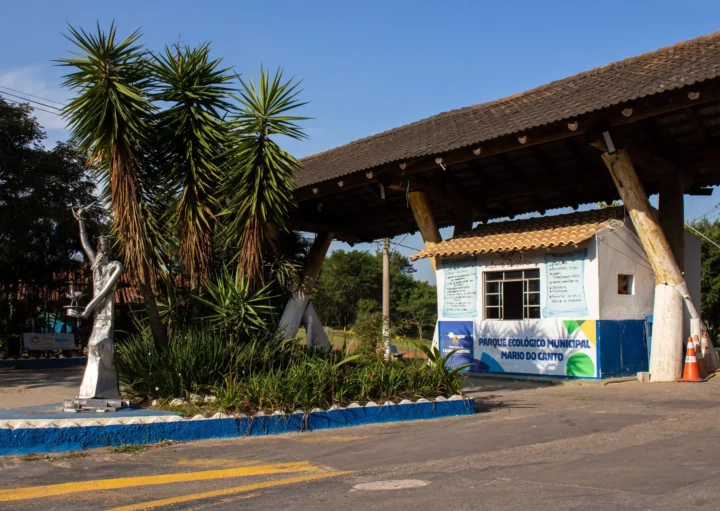 Portão de entrada do Parque Mario do Canto, um dos charmosos parques em Itaquaquecetuba, com um pequeno prédio branco e azul, placas de sinalização, plantas semelhantes a palmeiras, uma escultura de metal e árvores sob um céu ensolarado.