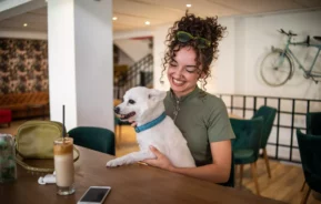 Uma mulher sorridente em um restaurante pet friendly sentada à mesa e com seu cachorro no colo