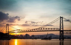 Imagem panorâmica da estrutura sobre o rio ao entardecer, com iluminação do sol, para ilustrar matéria sobre Ponte Hercílio Luz