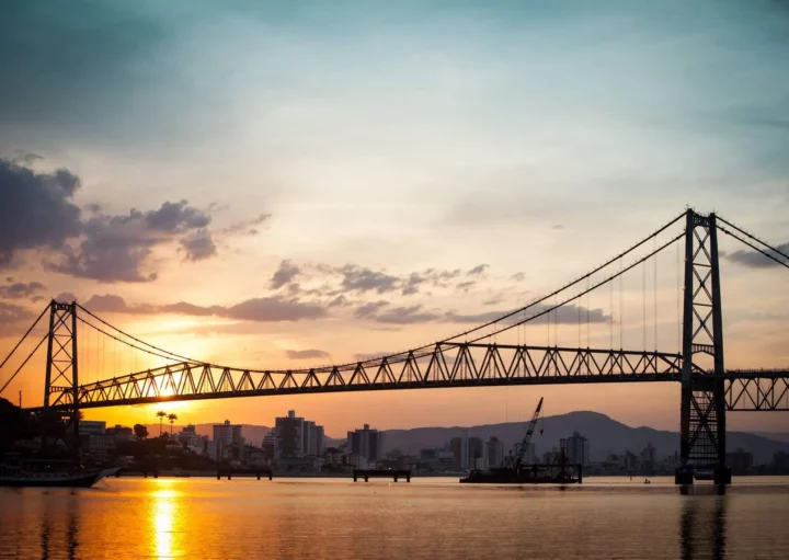Imagem panorâmica da estrutura sobre o rio ao entardecer, com iluminação do sol, para ilustrar matéria sobre Ponte Hercílio Luz