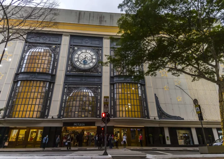 Imagem da fachada do shopping Pátio Paulista mostra relógio proeminente acima da entrada, decorado com estrelas douradas. As pessoas ficam na faixa de pedestres em frente à loja e as árvores altas emolduram parcialmente o edifício