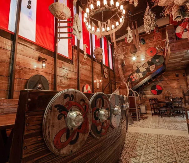 O interior do bar Taverma Medieval decorado com tema viking, apresentando uma réplica de navio viking de madeira com escudos pintados, paredes de madeira, faixas vermelhas e brancas, escudos pendurados e um lustre de vela. É um dos Bares temáticos em São Paulo