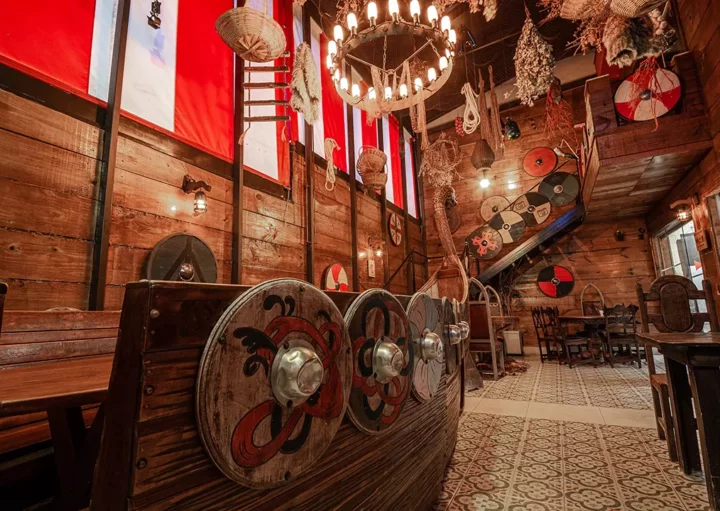 O interior do bar Taverma Medieval decorado com tema viking, apresentando uma réplica de navio viking de madeira com escudos pintados, paredes de madeira, faixas vermelhas e brancas, escudos pendurados e um lustre de vela. É um dos Bares temáticos em São Paulo