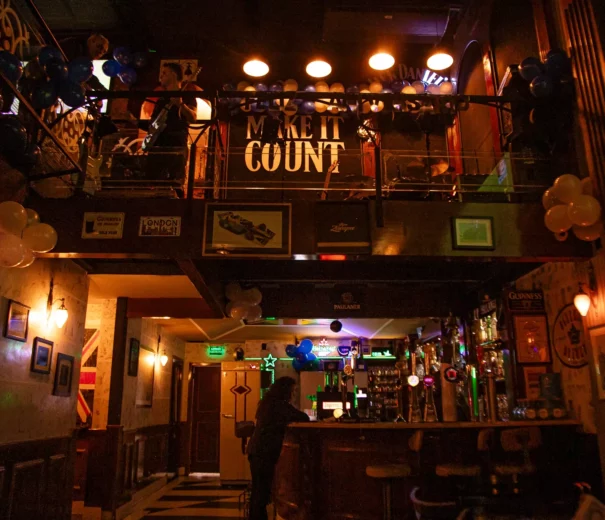Interior de um bar de rock em São Paulo, o The Blue Pub Itaim, com uma pessoa no balcão, balões para decoração e um balcão acima com uma grande placa onde se lê MAKE IT COUNT. Luzes quentes pendem do teto, criando uma atmosfera aconchegante.
