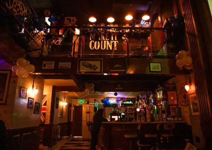 Interior de um bar de rock em São Paulo, o The Blue Pub Itaim, com uma pessoa no balcão, balões para decoração e um balcão acima com uma grande placa onde se lê MAKE IT COUNT. Luzes quentes pendem do teto, criando uma atmosfera aconchegante.