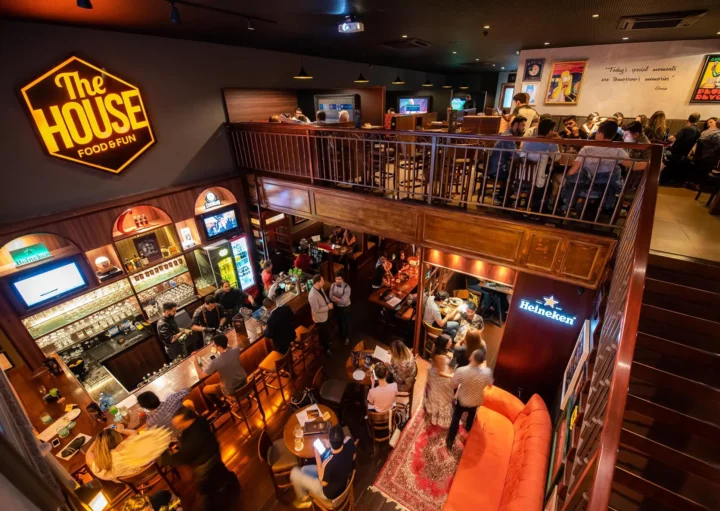 Foto mostrando a parte interna do The House Food & Fun com ângulo amplo podendo visualizar o salão no primeiro e segunda andar. O local é um bar temático em BH, parte dos dez mais famosos do segmento