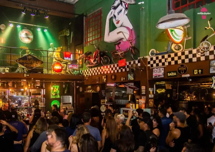 Um animado Bar de Rock em BH, o Underground Black Pub, com uma multidão assistindo a uma banda se apresentar em um palco na varanda, luzes coloridas, decoração vintage, um balcão de bar e um grande mural de uma mulher acima de motocicletas e letreiros de neon.