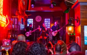 Uma banda se apresenta ao vivo no palco do Grainne’s Irish Pub, um bar de rock em Campinas, com uma cantora, um guitarrista, um baixista e um baterista. Uma multidão assiste, e luzes de neon vermelhas dão cor à cena.