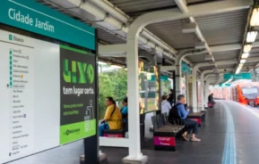 Pessoas sentadas e esperando em bancos na plataforma da estação Cidade Jardim do metrô de São Paulo; um trem laranja está chegando e uma placa promove o descarte adequado de resíduos.
