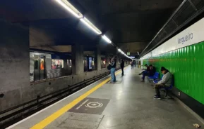 Uma plataforma de metrô com pessoas esperando, algumas sentadas em cadeiras azuis. Um trem azul está parado no trilho da esquerda. A parede da direita é verde e branca, exibindo o nome da estação Vergueiro. A iluminação é clara e uniforme.