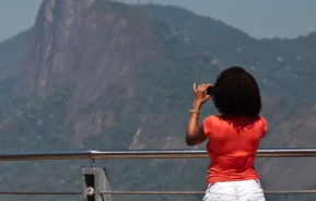 Foto que ilustra matéria sobre Mirantes no Rio de Janeiro mostra uma mulher de costas em uma mureta do Pão de Açúcar apontando uma câmera para tirar uma foto do Morro do Corcovado e do Cristo Redentor que aparece ao fundo (Foto: Getty Images)