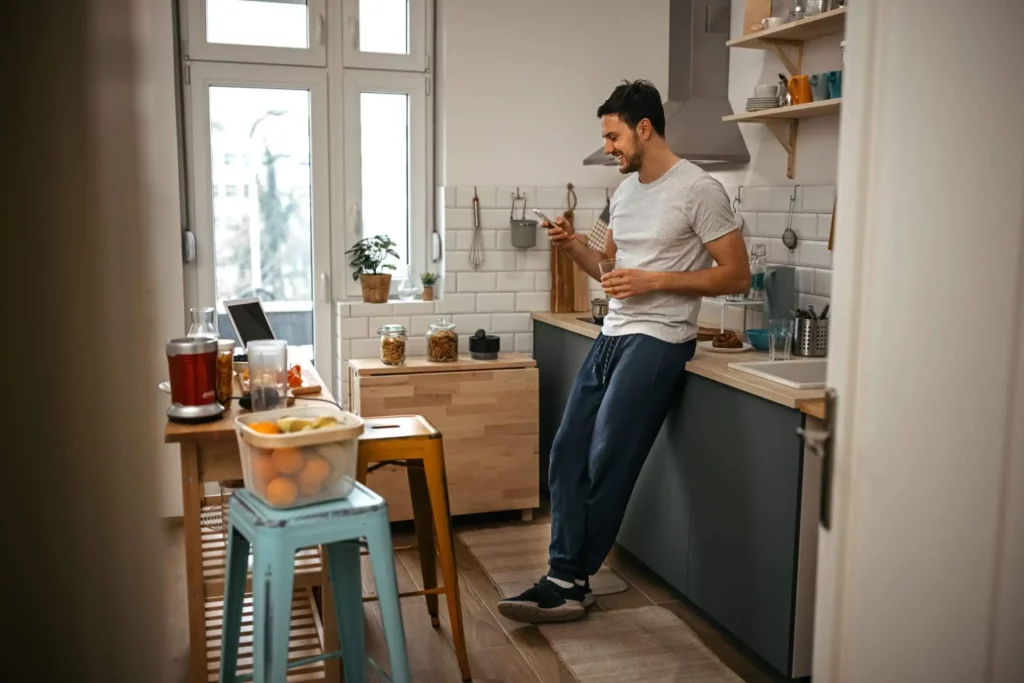 Imagem de um homem com roupas casuais em uma cozinha moderna, sorrindo para o telefone enquanto segura um copo. A bancada tem itens de cozinha e frutas, e a luz do sol entra pelas grandes janelas