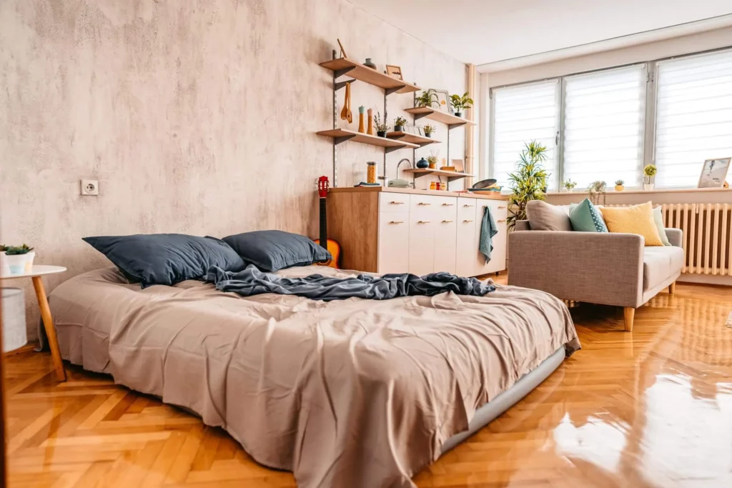Imagem de um apartamento aconchegante com uma cama não arrumada com travesseiros cinza e azul, um pequeno sofá, prateleiras de madeira com plantas e decoração, uma cozinha compacta e janelas grandes que permitem a entrada de luz natural