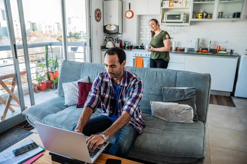 Imagem de um homem trabalhando em um notebook em uma mesa de centro em uma sala de estar bem iluminada, enquanto uma mulher está na cozinha atrás dele, segurando uma caneca. O espaço é aberto e moderno, com plantas e prédios da cidade visíveis através de grandes janelas
