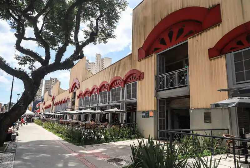 Imagem de uma ampla calçada ladeada por plantas verdes e assentos ao ar livre passa ao lado de um prédio do Mercado Municipal de Curitiba, com parede amarela, detalhes em arco vermelhos e grandes janelas. Uma árvore frondosa se ergue à esquerda sob um céu parcialmente nublado