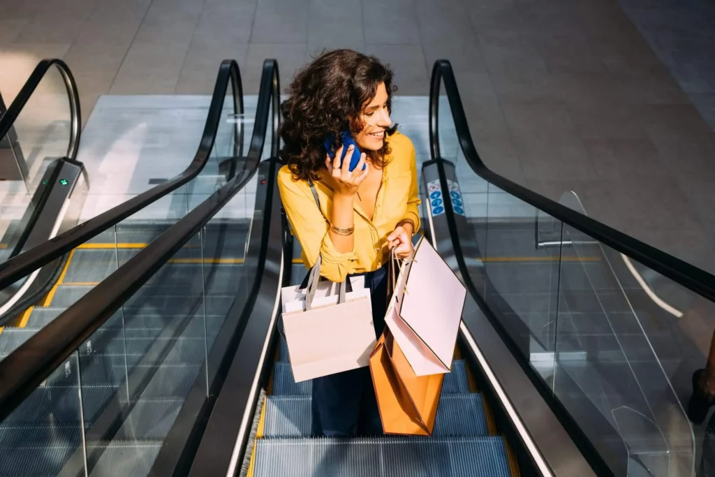 Imagem de uma mulher sorridente com cabelos cacheados, vestindo uma camisa amarela, está em uma escada rolante segurando sacolas de compras em uma mão e falando ao telefone com a outra, em um moderno shopping center