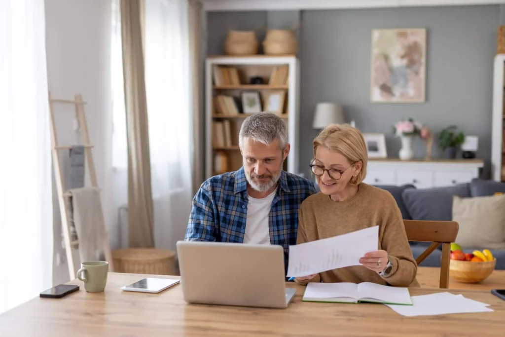 Imagem de um casal de idosos sentados junto a uma mesa, olhando para papéis e um notebook. Eles parecem estar analisando documentos, sorrindo e trabalhando juntos em uma sala de estar aconchegante e bem iluminada