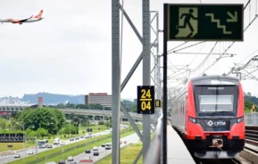 Um trem vermelho e preto do Expresso Aeroporto está parado em trilhos elevados. Abaixo, carros circulam em uma estrada movimentada. Um avião está voando no céu à esquerda. Há uma placa verde de saída de emergência e um relógio digital.
