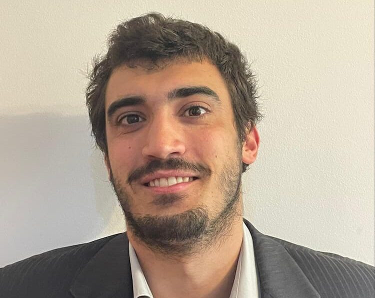 Um homem de cabelos escuros, barba curta e bigode está sorrindo levemente. Ele está vestindo um blazer escuro sobre uma camisa branca e está parado em frente a uma parede lisa e clara.