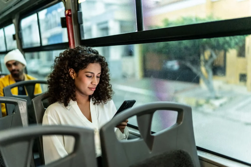 Imagem de uma mulher de cabelos cacheados sentada em um ônibus, olhando para seu smartphone. Ela está vestindo uma blusa branca. Do lado de fora da janela, uma rua embaçada e prédios são visíveis. Um homem com uma jaqueta amarela está sentado atrás dela