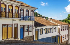 Imagem de casas coloridas em Ouro Preto, em Minas Gerais, em estilo colonial com telhados de telha e portas de madeira se alinham em uma rua íngreme de paralelepípedos sob um céu azul, com árvores e casas distantes visíveis ao fundo
